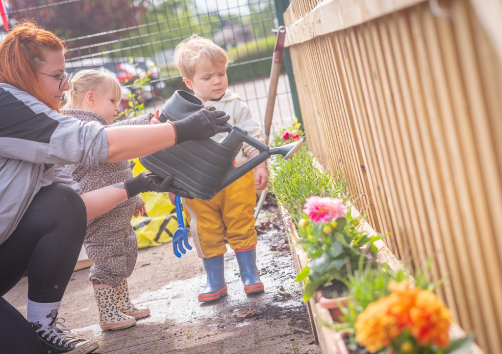 A team member from Cosy Direct helps children from the Little Bears room to water the new freshly panted flower tubs, as they watch on in awe.